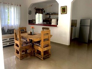 A dining area with a wooden table and chairs, a small kitchen area with a refrigerator, and a window with curtains. - Family Suite - Apartment 1 in Villa Coconut (Samaná)