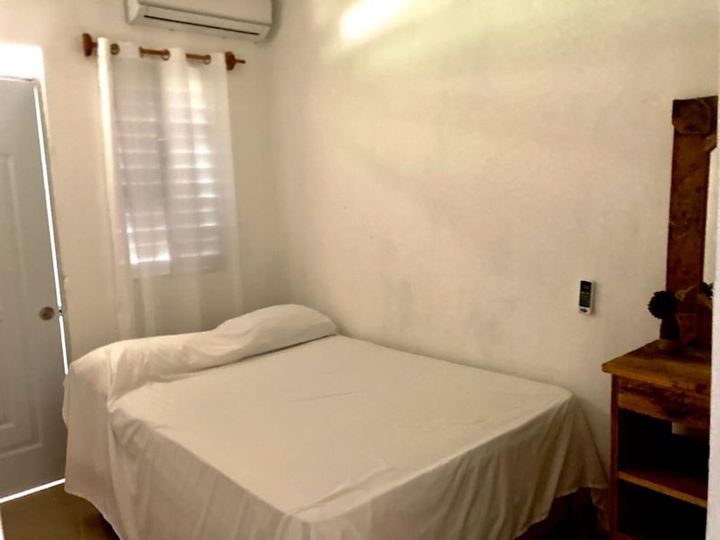 A small, single-bed room with a white bedspread, a wooden nightstand, a mirror, and a window with sheer curtains.