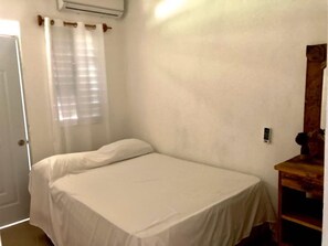 A small, single-bed room with a white bedspread, a wooden nightstand, a mirror, and a window with sheer curtains. - Family Suite - Apartment 1 in Villa Coconut (Samaná)