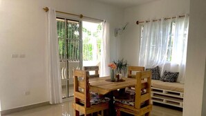 A dining area with a wooden table and chairs, a sofa with cushions, and a window with curtains. - Family Suite - Apartment 1 in Villa Coconut (Samaná)