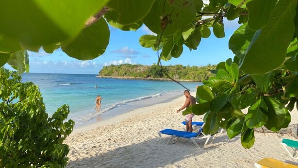 A beach with clear turquoise water, a sandy shore, and a person sunbathing on a blue lounge chair. - Family Suite - Apartment 1 in Villa Coconut (Samaná)