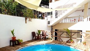 Ein Schwimmbecken mit Holzterrasse, eine Sitzgelegenheit mit Stühlen und einem Tisch, und ein weißes Gebäude mit Balkonen im Hintergrund.