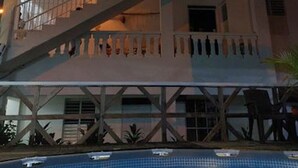 Ein zweigeschossiges Gebäude mit einem Balkon im oberen Stockwerk, einer Treppe, die zum Balkon führt, und einem Schwimmbecken im Vordergrund.