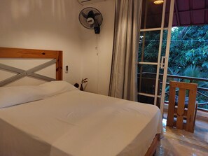 A bedroom with a wooden headboard, a bed with white linens, a wall-mounted fan, a window with curtains, and a view of greenery outside. - Family Apartment - Apartment 5 in Villa Coconut (Samaná)