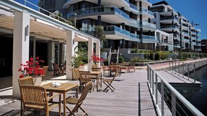 A waterfront outdoor seating area with wooden tables and chairs, a covered walkway, and a modern building with balconies in the background.