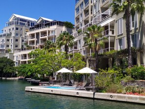 Un hotel frente al agua con varios balcones, una zona de piscina con tumbonas y abundante vegetación.