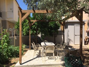 A covered outdoor seating area with a table and chairs, surrounded by greenery and a pergola structure. - Room in Studio - Beautiful Room in Limenaria, Only Five Minutes Away From Center (Thasos)