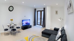 A modern living room with a dining area, a flat-screen TV, a clock, and a balcony. - Alexandra Palace Luxury Serviced Apartments In St Albans (St Albans)