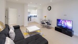 A modern living room with a sofa, a coffee table, a dining table with chairs, a TV, and a kitchen in the background. - Alexandra Palace Luxury Serviced Apartments In St Albans (St Albans)