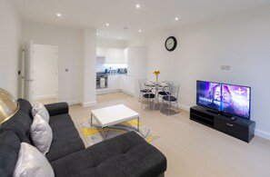 Un salon moderne avec un canapé, une table basse, une table à manger avec des chaises, un téléviseur et une cuisine en arrière-plan.