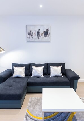 A modern living room with a dark grey sectional sofa, three patterned throw pillows, a white coffee table, and a framed abstract painting of horses on the wall. - Alexandra Palace Luxury Serviced Apartments In St Albans (St Albans)