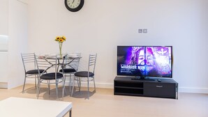 A modern living room with a dining table, chairs, a wall clock, and a TV displaying a show. - Alexandra Palace Luxury Serviced Apartments In St Albans (St Albans)