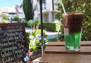 Ein Glas Eistee mit Trinkhalm auf einem Holztisch, eine Tafelnmenü mit Getränken und Happy-Hour-Angeboten sowie ein Gebäude mit Balkonen und Bepflanzung im Hintergrund.