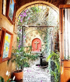 An arched doorway leading to a courtyard with a fountain, surrounded by potted plants and a red door.
