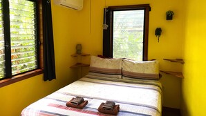 A bedroom with a bed, two pillows, a bedside table with a lamp, a window with blinds, and an air conditioning unit. - Private 2-bedroom Jungle Cabin in Tropical Garden, San Ignacio (San Ignacio)