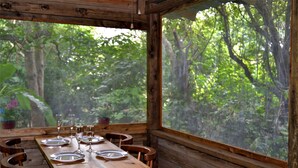 A wooden dining table set with plates, glasses, and cutlery, surrounded by chairs, with a large window offering a view of dense greenery. - Private 2-bedroom Jungle Cabin in Tropical Garden, San Ignacio (San Ignacio)
