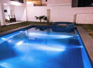 Zwembad met jacuzzi, omringd door een wit gebouw met een deur en een kleine palmboom.