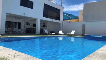 Un área de piscina moderna al aire libre, con una piscina de color azul cristalino, sillas de descanso blancas y una zona de estar cubierta.