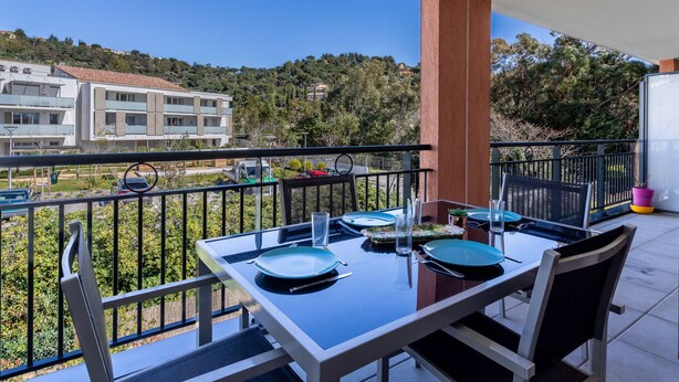A balcony with a table set for dining, overlooking a residential area with buildings and trees. - Le Jardin du Loderi by Interhome (Bormes-les-Mimosas)