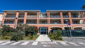 A multi-story hotel with a central entrance, balconies on each floor, and a parking area in front. - Le Jardin du Loderi by Interhome (Bormes-les-Mimosas)