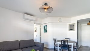 A modern living room with a grey sofa, a wooden coffee table with a plant, a dining area with a white table and dark chairs, and a wall clock. - Le Jardin du Loderi by Interhome (Bormes-les-Mimosas)