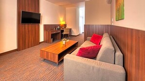 A hotel room with a sofa, a coffee table, a TV mounted on the wall, a desk with a chair, and a lamp. - Centrum (Kosice)