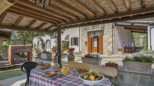 An outdoor patio with a brick oven, a table set with bowls and fruits, and a stone building in the background. - Cosy apartment in villa for 3 guests with pool, WIFI, TV and patio (Castelfranco Di Sopra)
