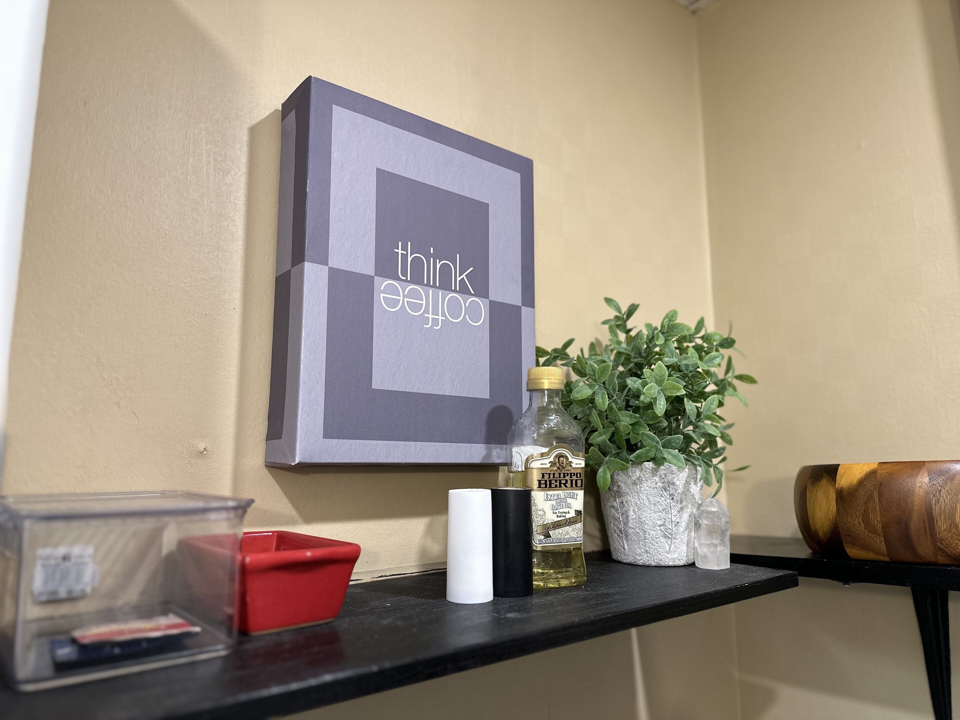 Une étagère avec une œuvre d’art encadrée, une bouteille d’huile d’olive extra légère de la marque Filippo, une bougie blanche, un récipient rouge et une plante en pot.