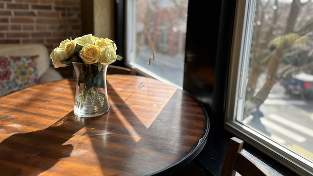 Une table ronde en bois sur laquelle est posé un vase en verre transparent contenant des roses jaunes. La table est placée près d’une fenêtre donnant sur une rue et des voitures garées.