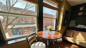 Une pièce chaleureuse avec une table à manger en bois, deux chaises, un vase rempli de fleurs jaunes, un canapé agrémenté d’un coussin à motifs et une vue sur un bâtiment en briques à l’extérieur.
