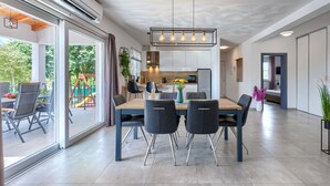 Una cocina moderna con una isla central, un comedor con una mesa grande y sillas, y vistas a un jardín a través de ventanas amplias.