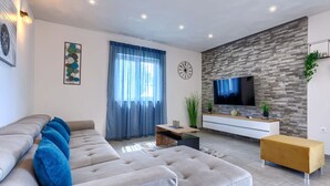 Una sala de estar moderna con un sofá seccional gris, un taburete amarillo, un televisor montado en una pared con textura y un ventanal con cortinas azules.