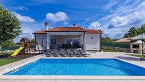 Una piscina con un porche cubierto, una estructura de juegos para niños y un muro de piedra.