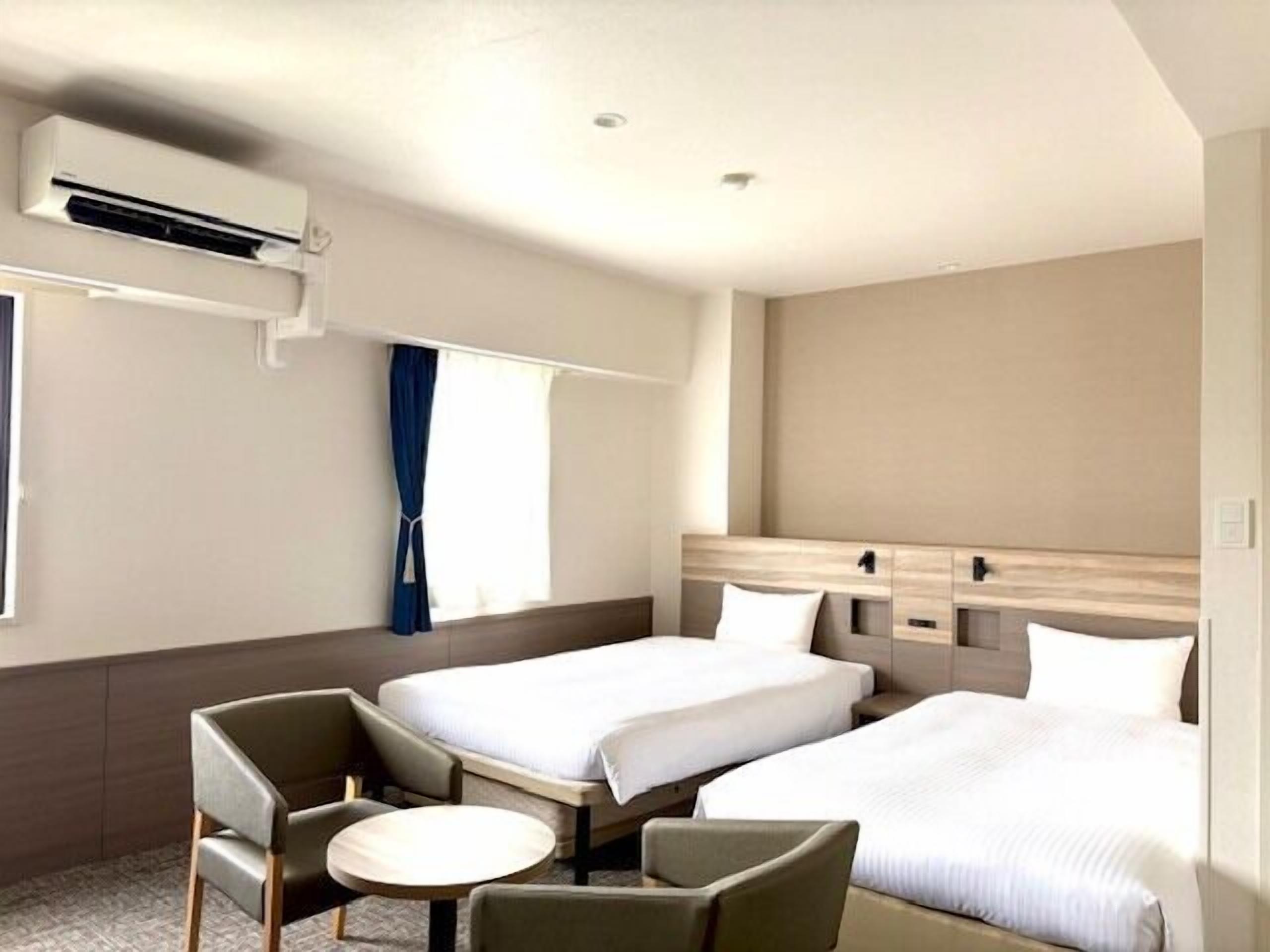 Hotelkamer met twee bedden, een kleine tafel, een stoel, een airconditioner en een raam met gordijnen.