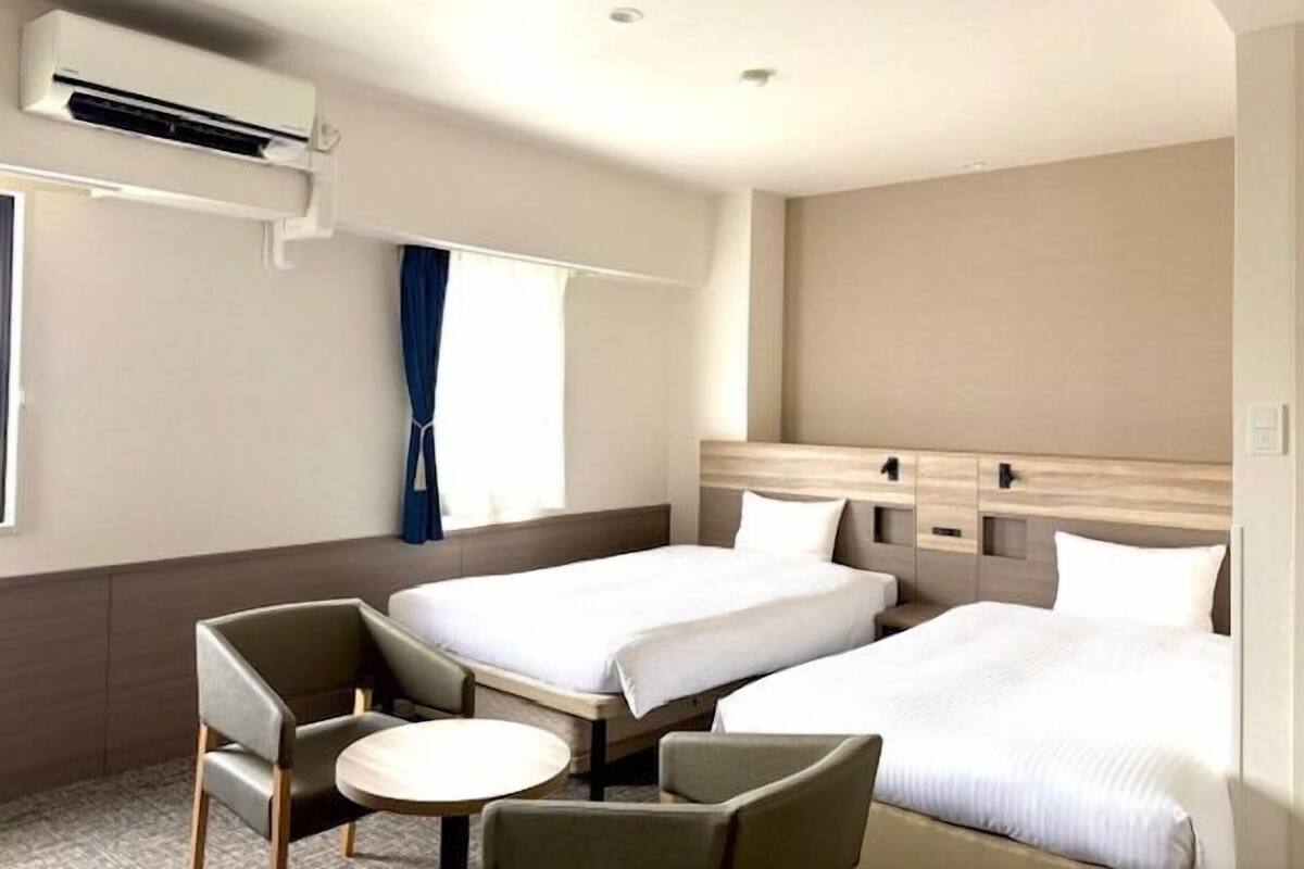 Habitación de hotel con dos camas, una mesita, una silla, un equipo de aire acondicionado y una ventana con cortinas.