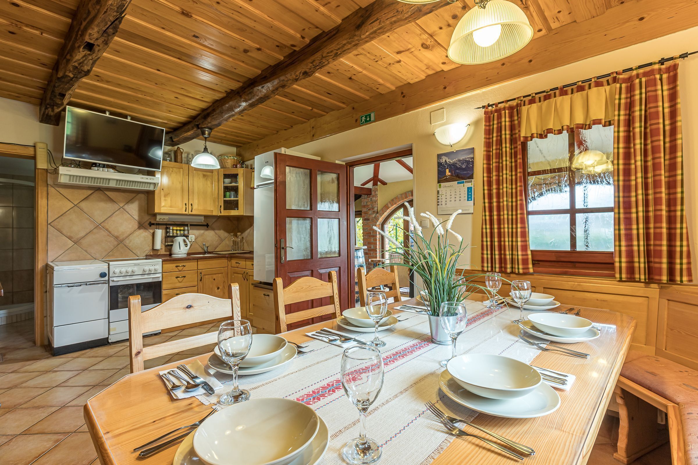 Une table de salle à manger en bois, mise avec des assiettes, des verres et des couverts, une cuisine avec des meubles et des appareils électroménagers en bois, et une fenêtre ornée de rideaux à carreaux.