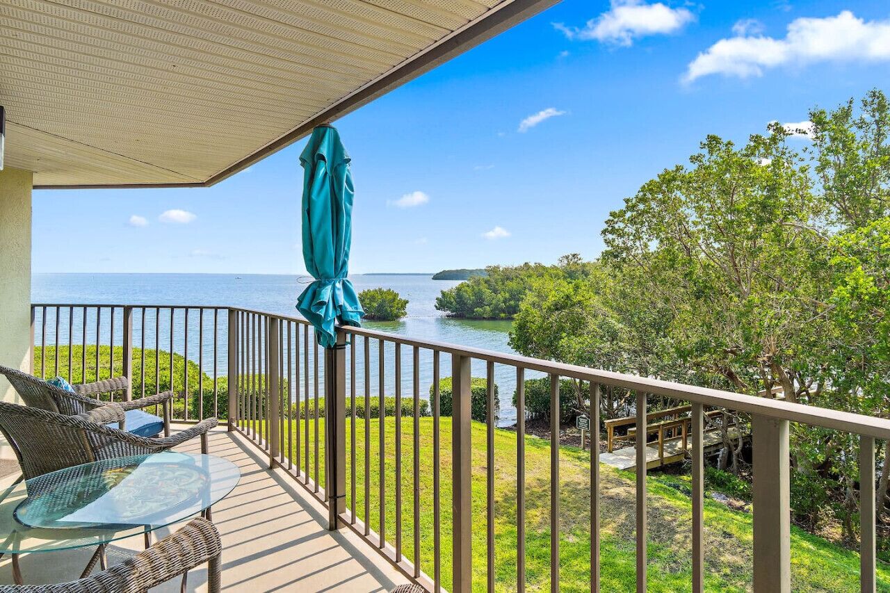 A balcony with wicker furniture and a glass table, overlooking a body of water and greenery.