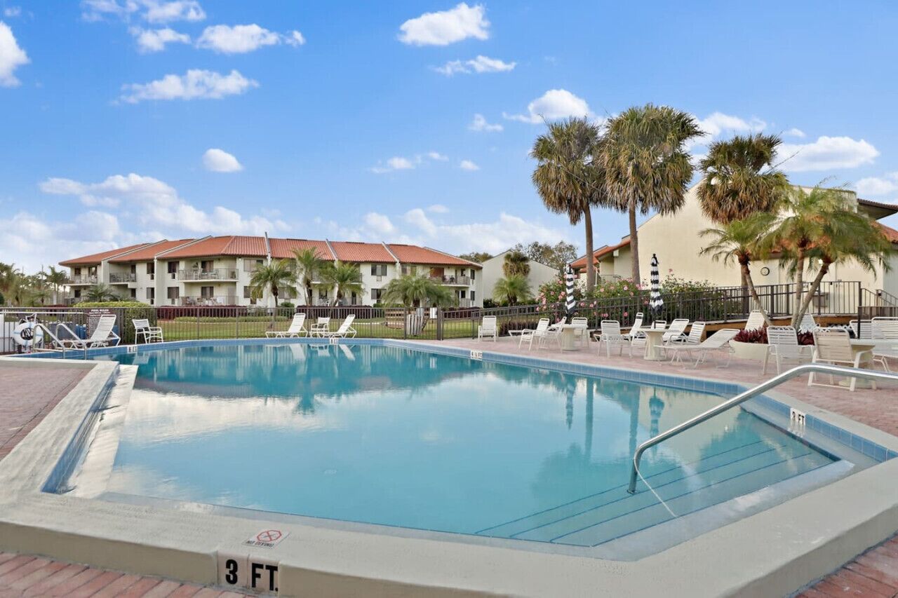 A swimming pool with lounge chairs and palm trees, surrounded by buildings with red roofs.