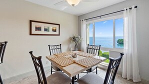 Un comedor con una mesa dispuesta para cuatro, un ventilador de techo y vistas al océano a través de amplias ventanas.