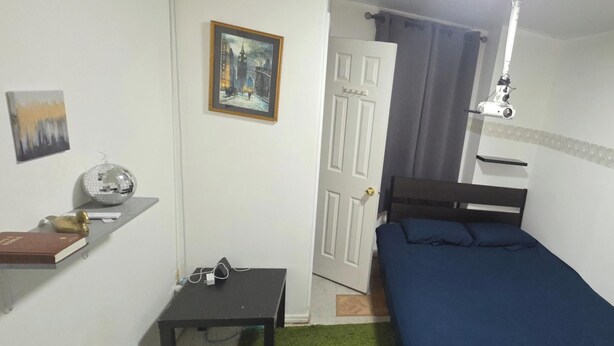 A small room with a bed, a desk, a chair, a shelf with books and a decorative item, and a door leading to another room. - Private Suite & Bath  10 min JFK  Flexible Terms (Richmond Hill)