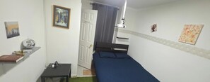 Een kleine kamer met een bed, een bureau, een stoel, een plank met boeken en een decoratief object, en een deur naar een andere kamer.