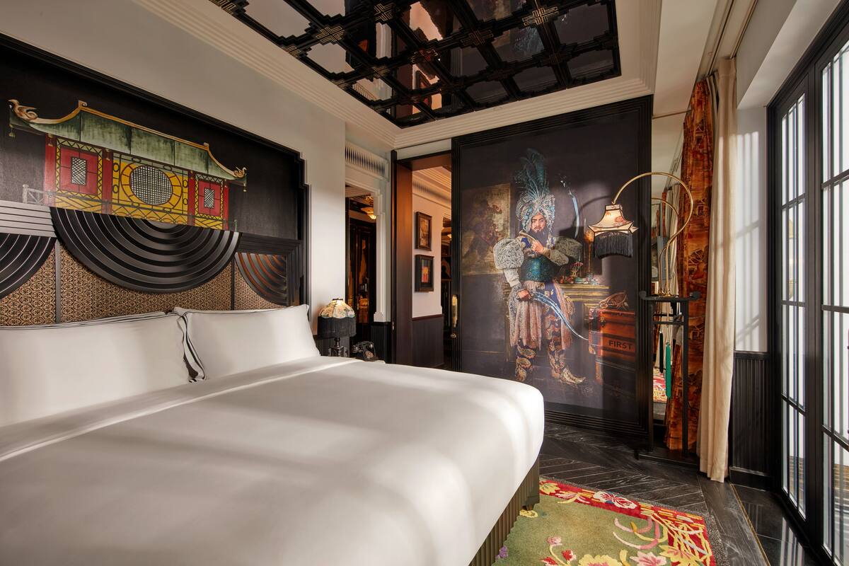 A modern hotel room with a large bed, a decorative headboard, a traditional Chinese mural, and a view of the city through large windows.
