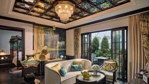 A spacious living room with a large sofa, a coffee table, and a chandelier. There is a painting on the wall and a window with a view of trees. - Capella Hanoi (Hanoi)