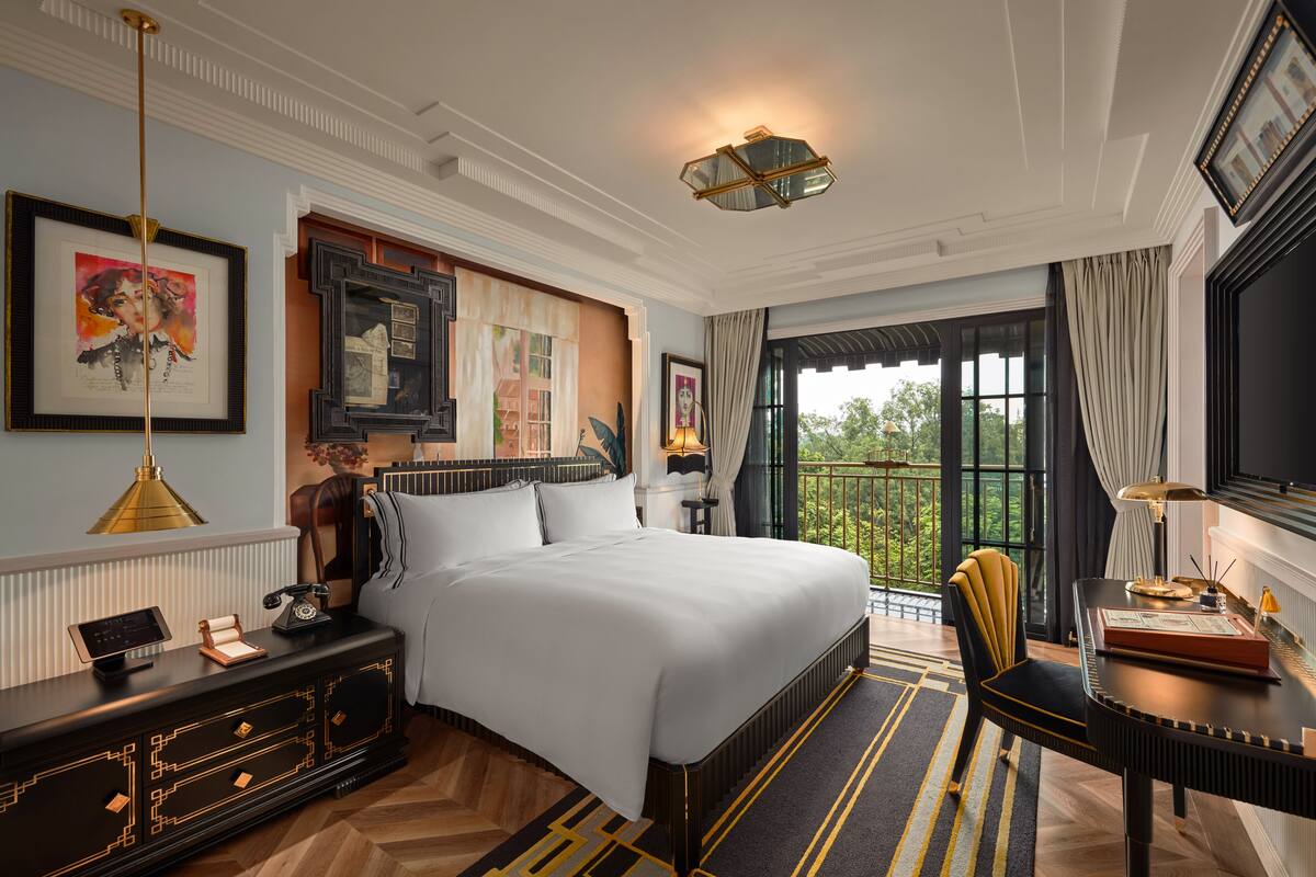 A spacious bedroom with a large bed, a desk with a lamp, a chair, and a view of the outdoors.