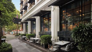 An outdoor cafe with tables and chairs, large windows, and greenery. - Capella Hanoi (Hanoi)