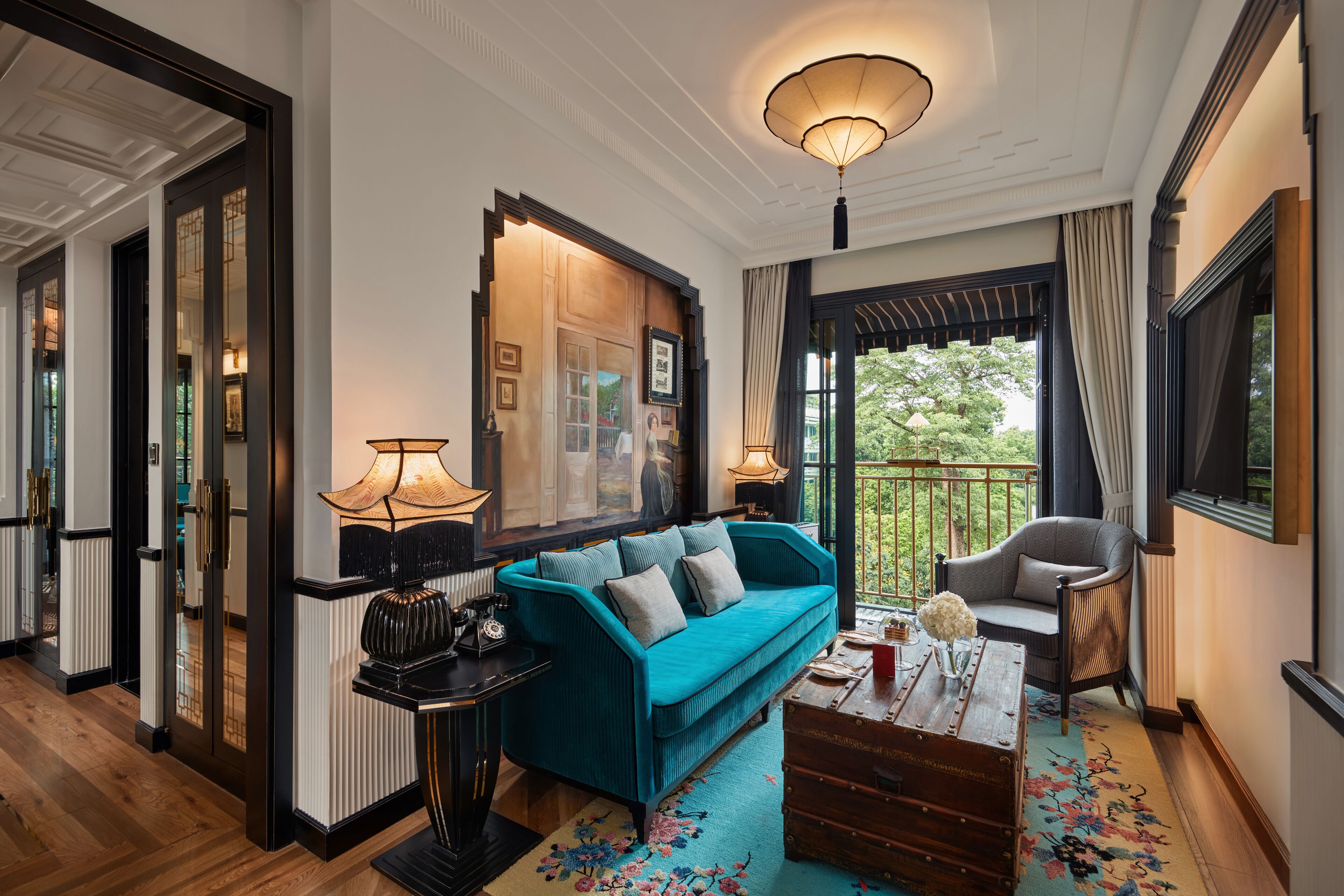 A room with a teal sofa, a wooden chest of drawers, a black lamp, and a large window with a view of greenery.