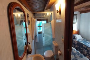 Baño pequeño con inodoro, lavamanos y ducha. Se ve una cama con ropa de cama estampada en la habitación contigua.