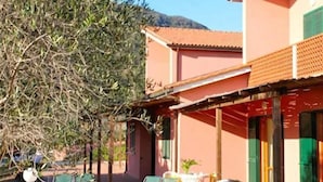 Un edificio con fachada roja y contraventanas verdes, con mesas y sillas en la terraza, y un árbol en primer plano.