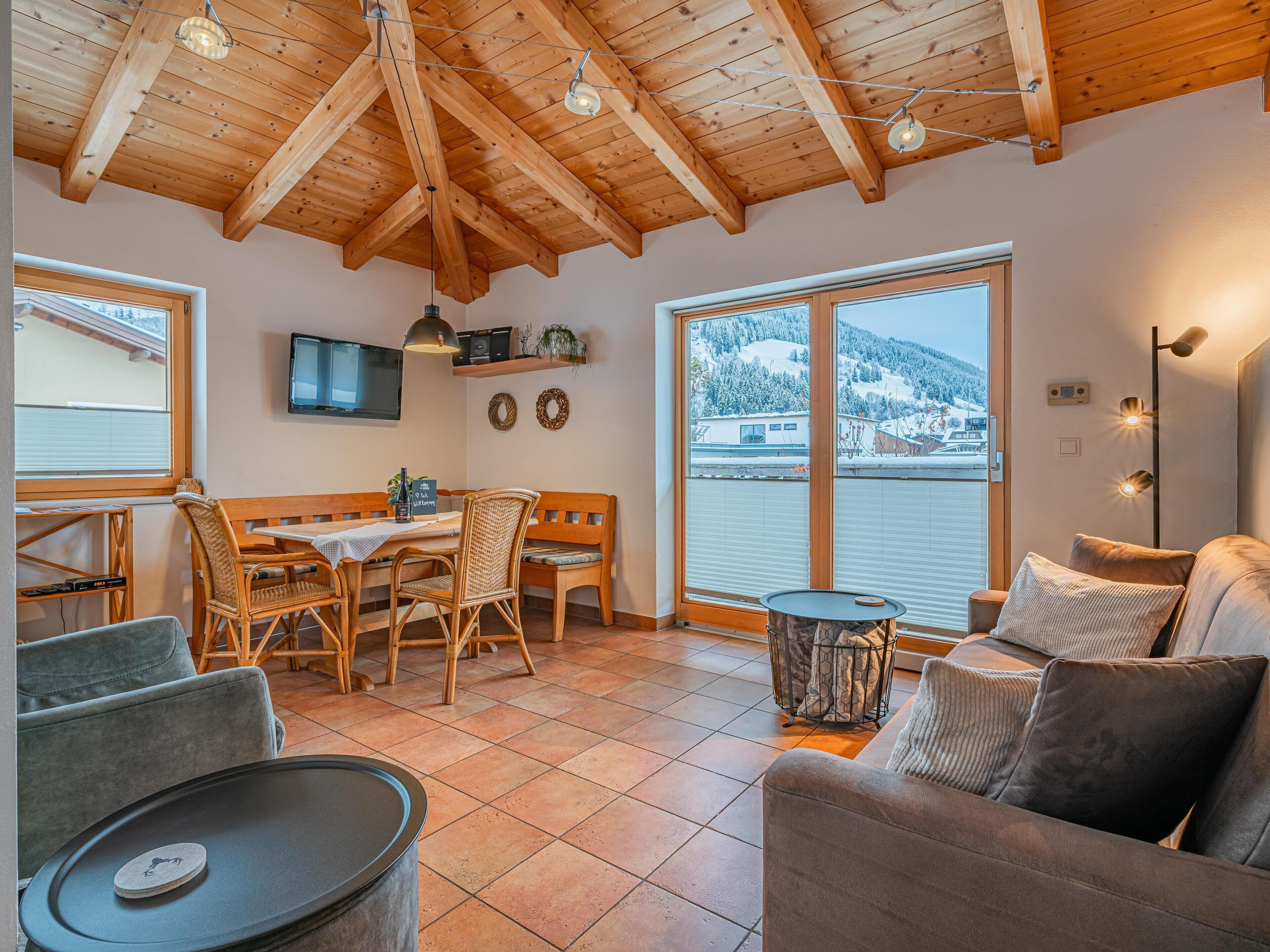 Ein gemütliches Zimmer mit Holzbalkendecke, einem Essbereich mit Tisch und Stühlen, einer Couch und einem großen Fenster mit Blick auf eine verschneite Landschaft.
