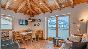 A cozy room with a wooden ceiling, a dining area with a table and chairs, a sofa, and a large window offering a view of a snowy landscape. - Detached holiday home in centre of Mittersill (Mittersill)